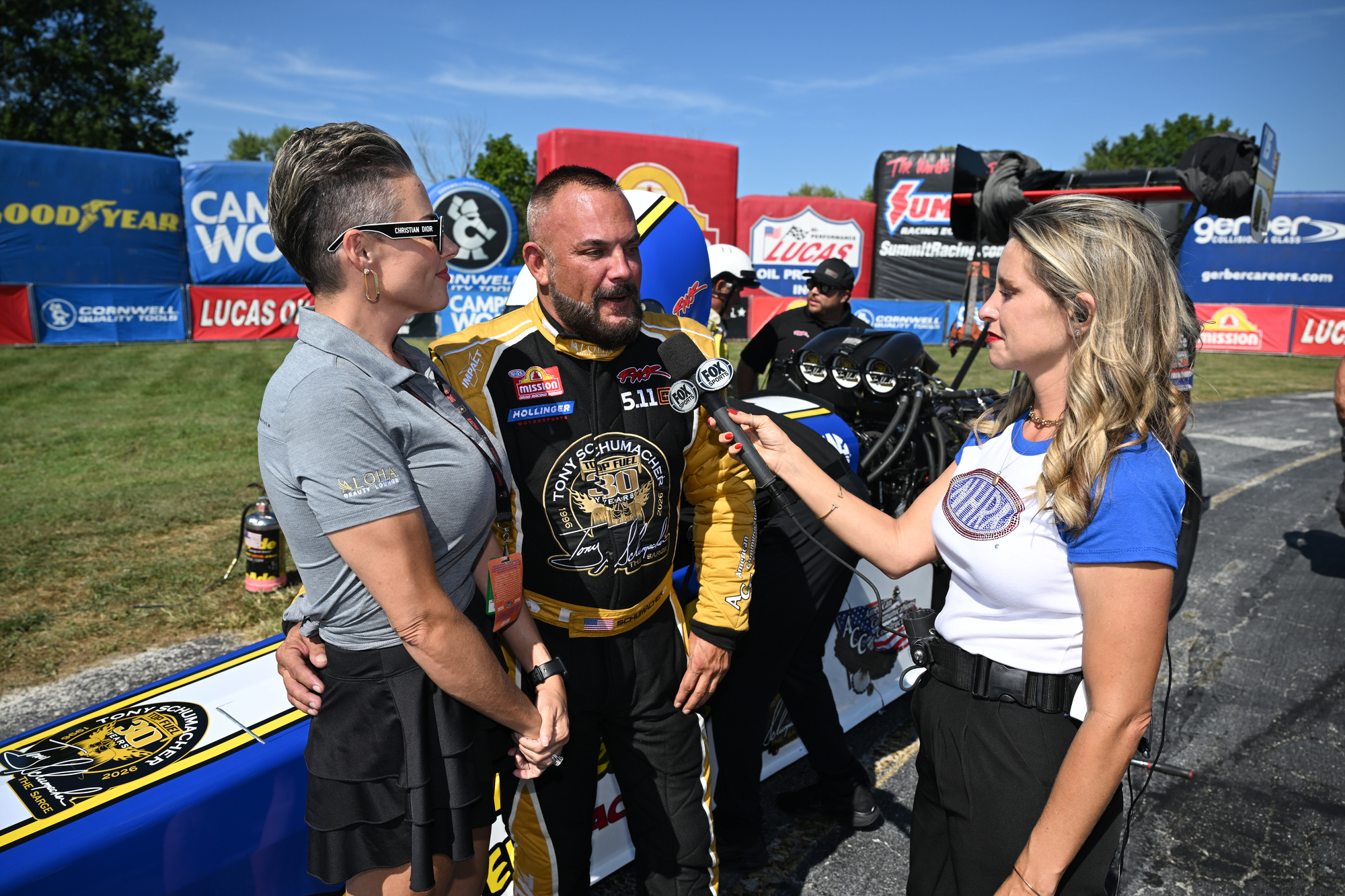 Tony Schumacher is interviewed beside his dragster in the pits at Indianapolis wearing the black and gold 30 Years tribute firesuit