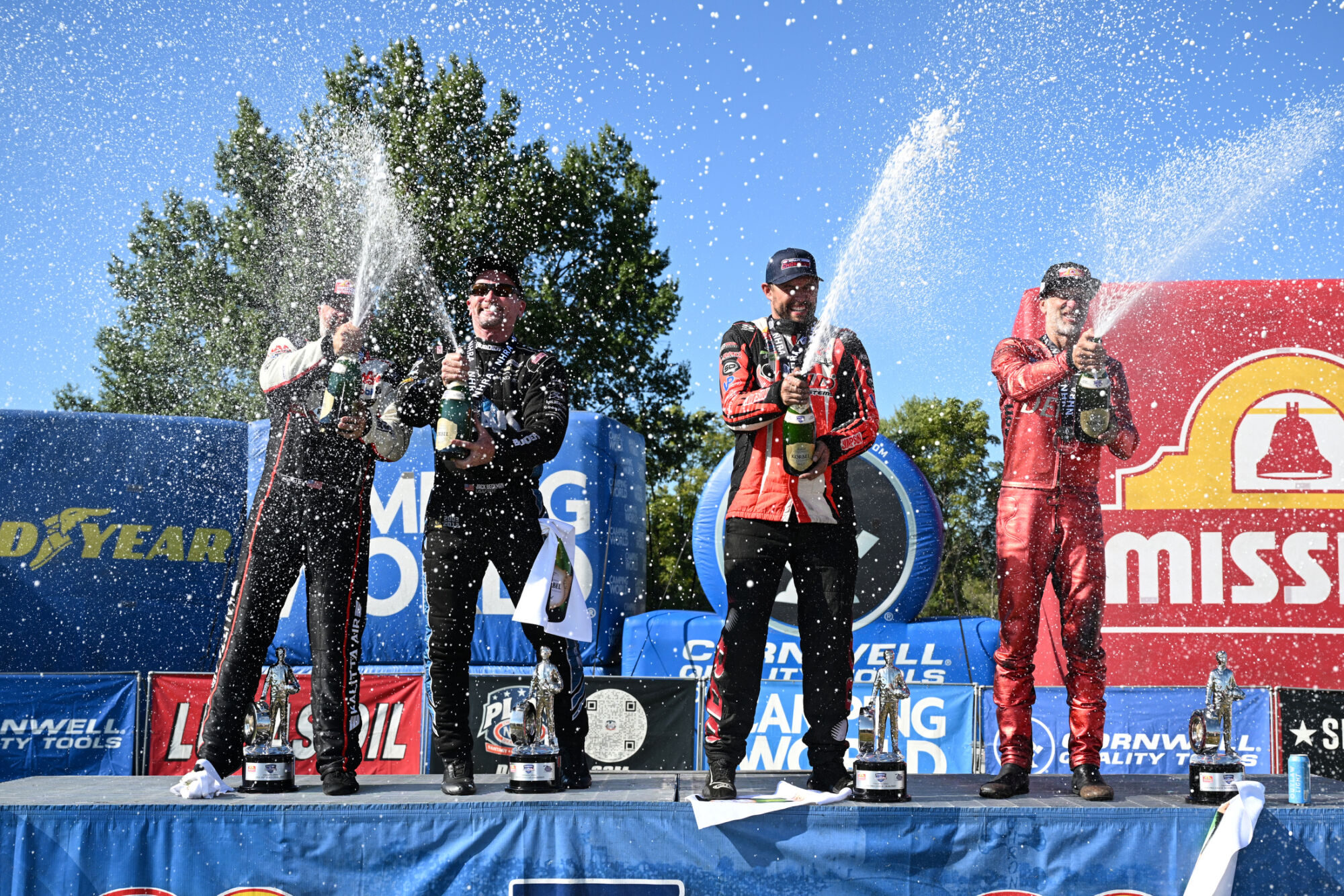 The four professional winners spray champagne on the winners stand at the 2026 NHRA U.S. Nationals