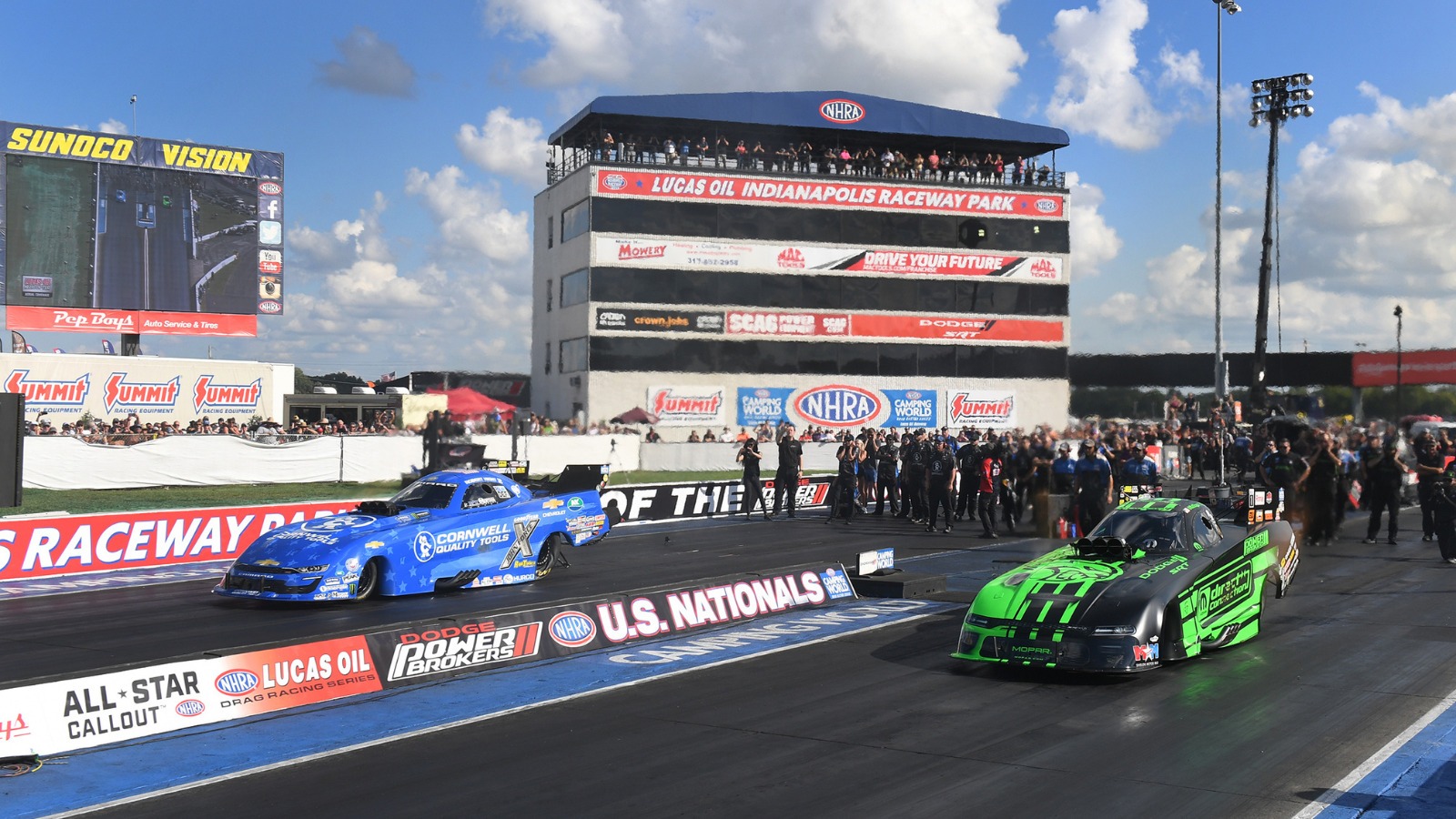 Two nitro Funny Cars leave the starting line side by side at the NHRA U.S. Nationals with the Lucas Oil Indianapolis Raceway Park tower behind them
