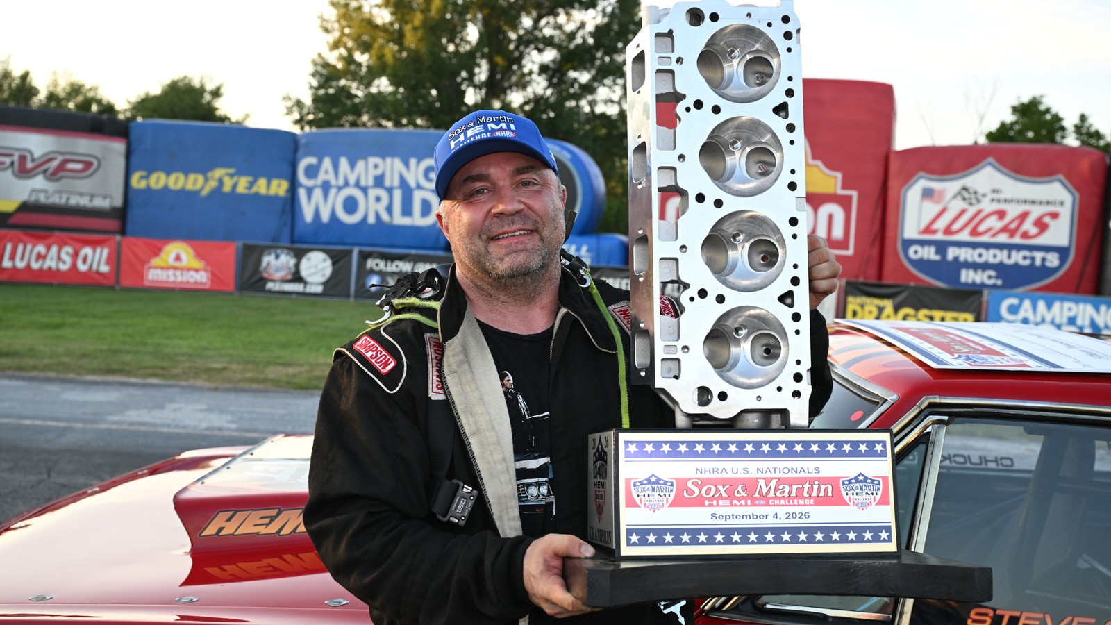 Steve Comella holds the Sox and Martin Hemi Challenge winner trophy, a Hemi cylinder head on a wooden base, beside his red 1968 Barracuda