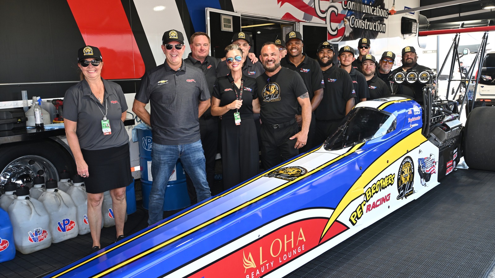 Tony Schumacher and the Rick Ware Racing team stand behind the blue, yellow and white Peek Brothers tribute Top Fuel dragster in the pits at Indianapolis