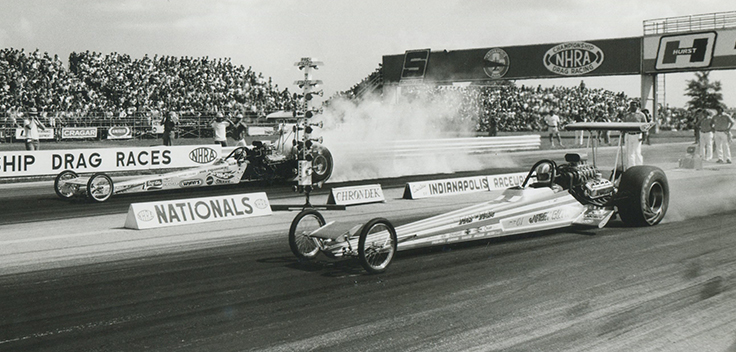 Black and white photo of two front engine dragsters launching side by side at Indianapolis Raceway Park during the U.S. Nationals, packed grandstands behind